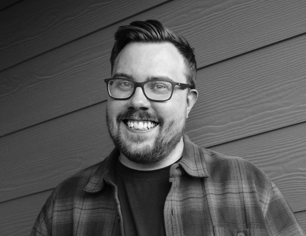 Black and white portrait of Andrew Adams, smiling, in glasses and a plaid shirt.